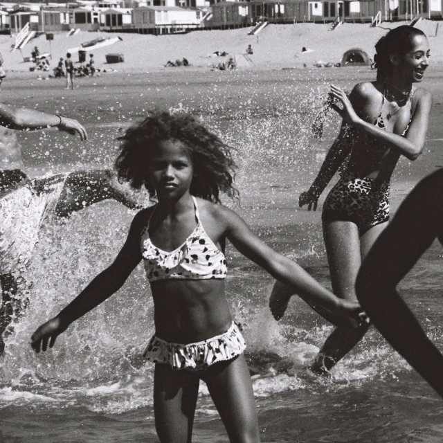 Black and white photograph of a child in a polka-dot bikini walking through the water on a beach with adults and distant beach houses.