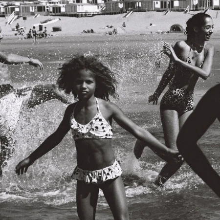 Black and white photograph of a child in a polka-dot bikini walking through the water on a beach with adults and distant beach houses.