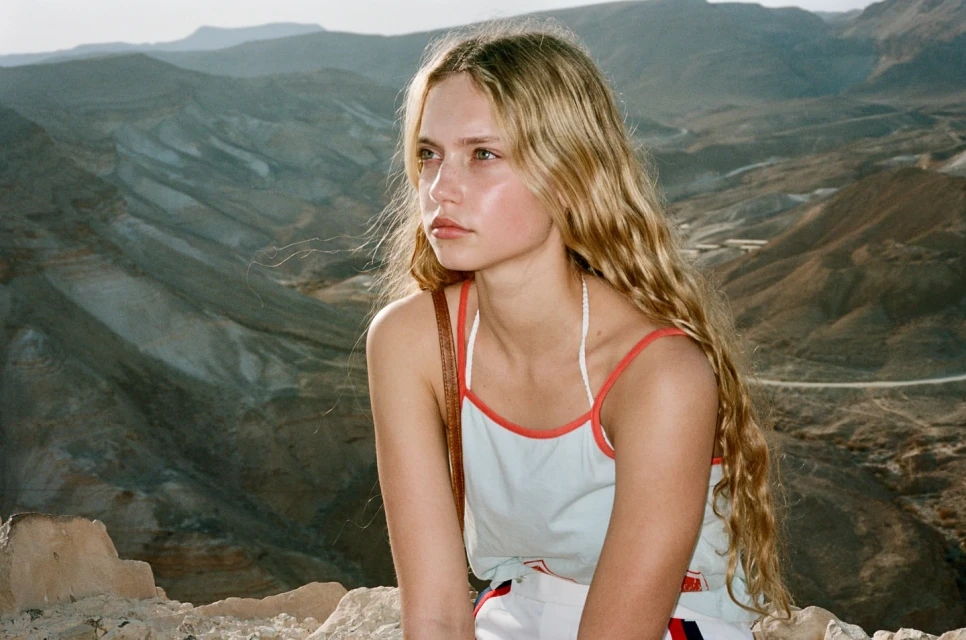 Portrait of a blonde woman with long wavy hair looking towards mountains on a sunny day, wearing a light blue tank top.