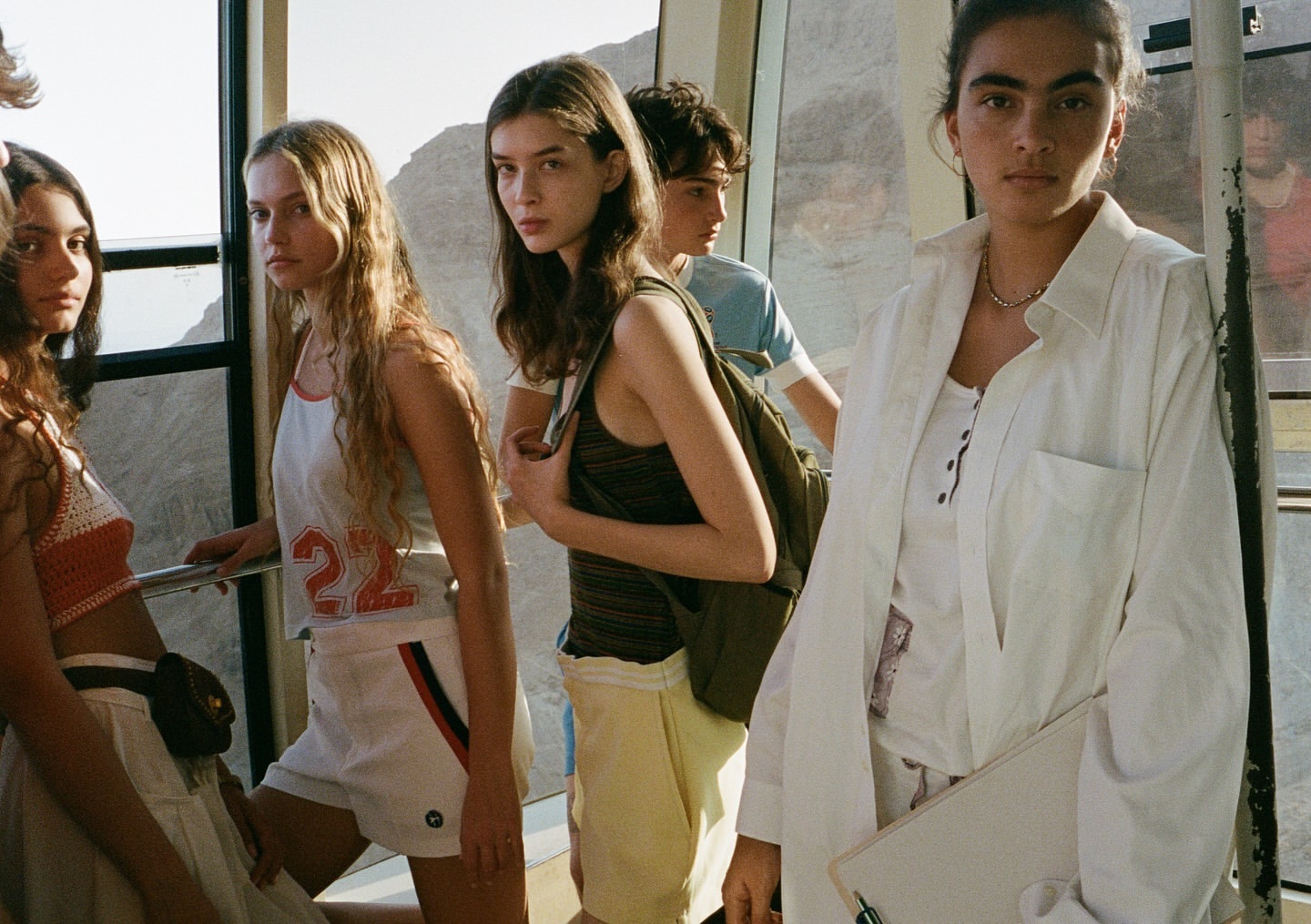 Group of young adults in a cable car, wearing casual summer clothing, with mountains visible through the windows in the background.