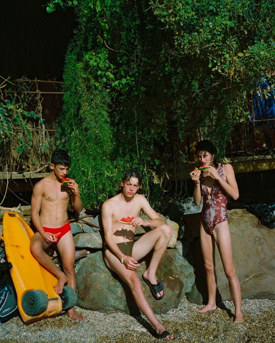Three young adults, two males and one female, sit on a rock formation outside, eating watermelon, in a natural setting with lush foliage.