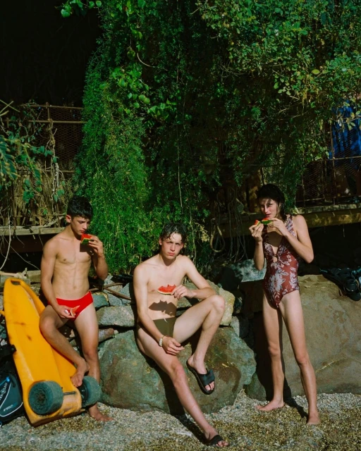 Three young adults, two males and one female, sit on a rock formation outside, eating watermelon, in a natural setting with lush foliage.