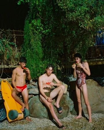 Three young adults, two males and one female, sit on a rock formation outside, eating watermelon, in a natural setting with lush foliage.