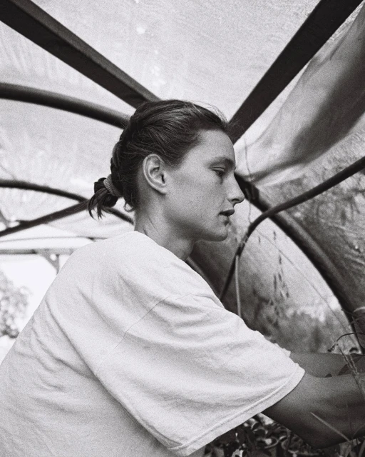 Woman tending to plants inside of a greenhouse, portrait view. The greenhouse has a translucent roof and the image is in black and white.