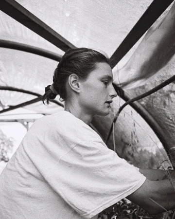 Woman tending to plants inside of a greenhouse, portrait view. The greenhouse has a translucent roof and the image is in black and white.