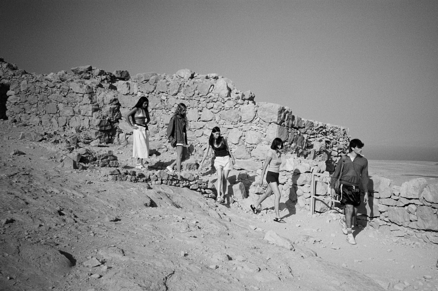 Group of six people walking down a sandy hill next to stone ruins in a black and white photograph under a clear sky.