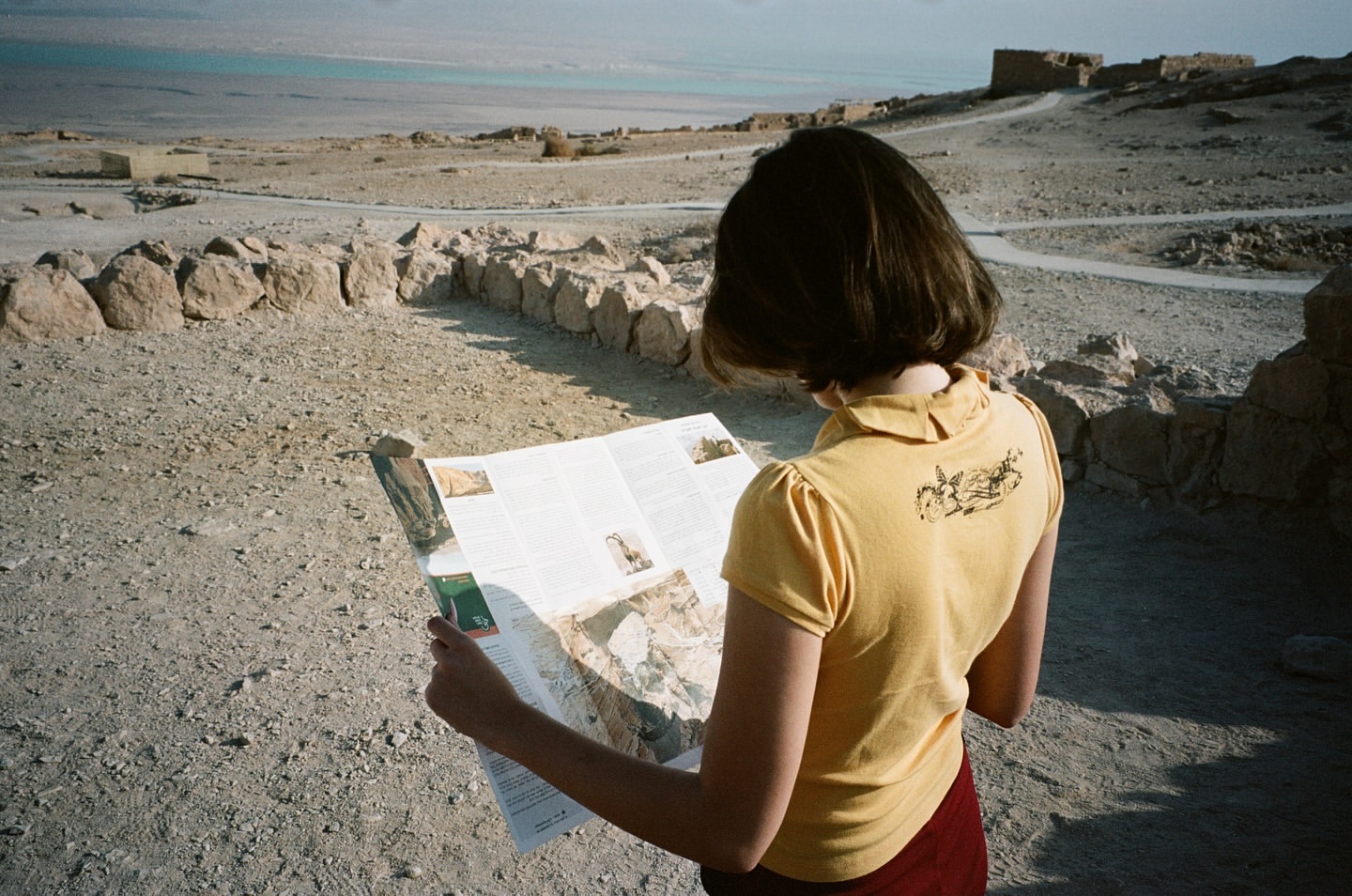 Woman in yellow shirt reads map at ancient ruins with arid landscape and sea view, creating a travel or historical scene in natural light.