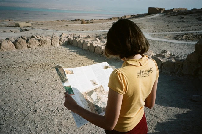 Woman in yellow shirt reads map at ancient ruins with arid landscape and sea view, creating a travel or historical scene in natural light.