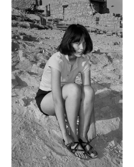 Monochrome portrait of a young woman in shorts and sandals, seated on a sandy slope with stone ruins in the background.
