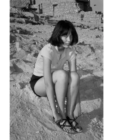 Monochrome portrait of a young woman in shorts and sandals, seated on a sandy slope with stone ruins in the background.