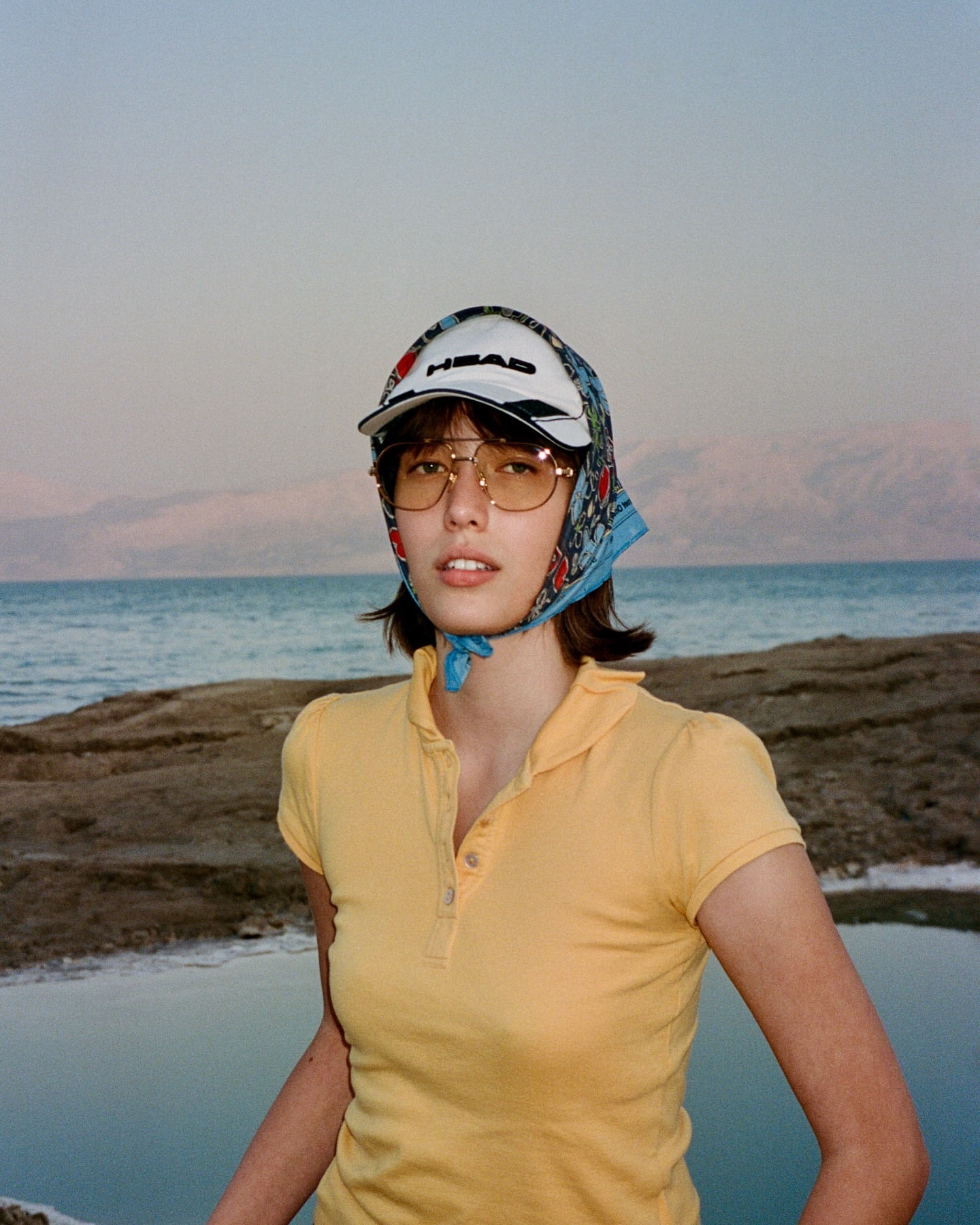 Young woman in a yellow polo shirt and floral scarf posing on a rocky beach with mountains and a pastel sunset sky in the background.
