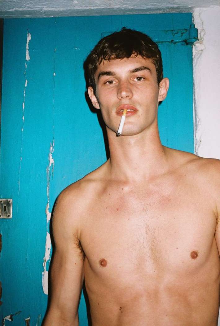 Shirtless man with short brown hair poses with a cigarette against a distressed blue wall in a medium studio portrait.