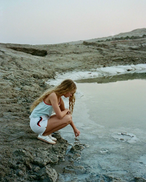Woman with blonde hair crouching by the water's edge, hand reaching toward the water in an arid, rocky landscape at dusk.