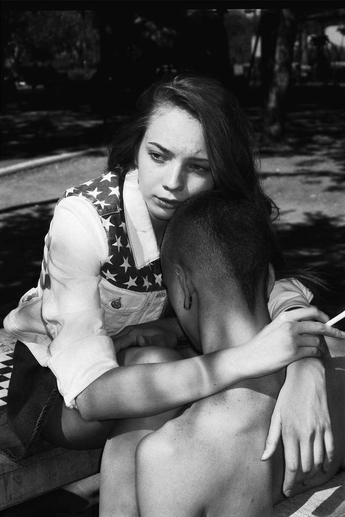 A young woman in a star-spangled vest embraces a shirtless person with a cigarette in an outdoor setting with trees in the background.