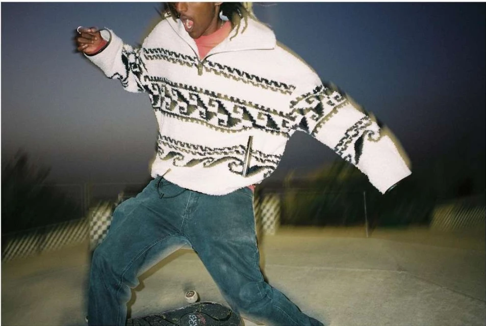 African American male wearing patterned sweater and jeans skateboarding on a concrete ramp at dusk; motion blur and shallow depth of field.