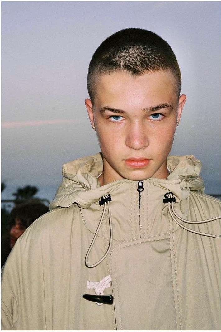 Close-up of a young adult male with a buzzcut wearing a beige hooded toggle coat, against a pale outdoor background.
