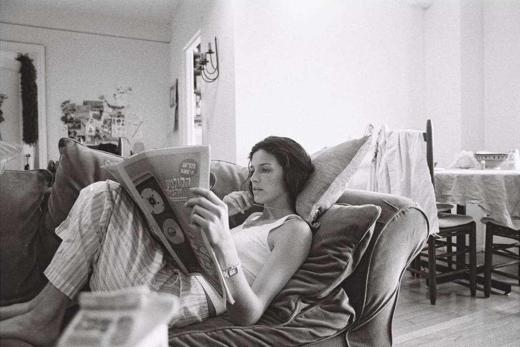 A woman in a white tank top and striped trousers relaxing on a plush sofa while reading a newspaper in an interior living space.