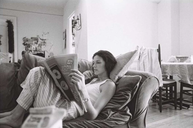 A woman in a white tank top and striped trousers relaxing on a plush sofa while reading a newspaper in an interior living space.