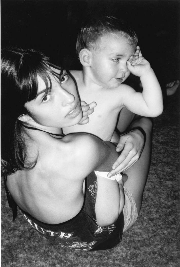 Black and white portrait of a young woman seated on the floor holding a toddler in her lap. The photo has a soft, fine-grain quality.