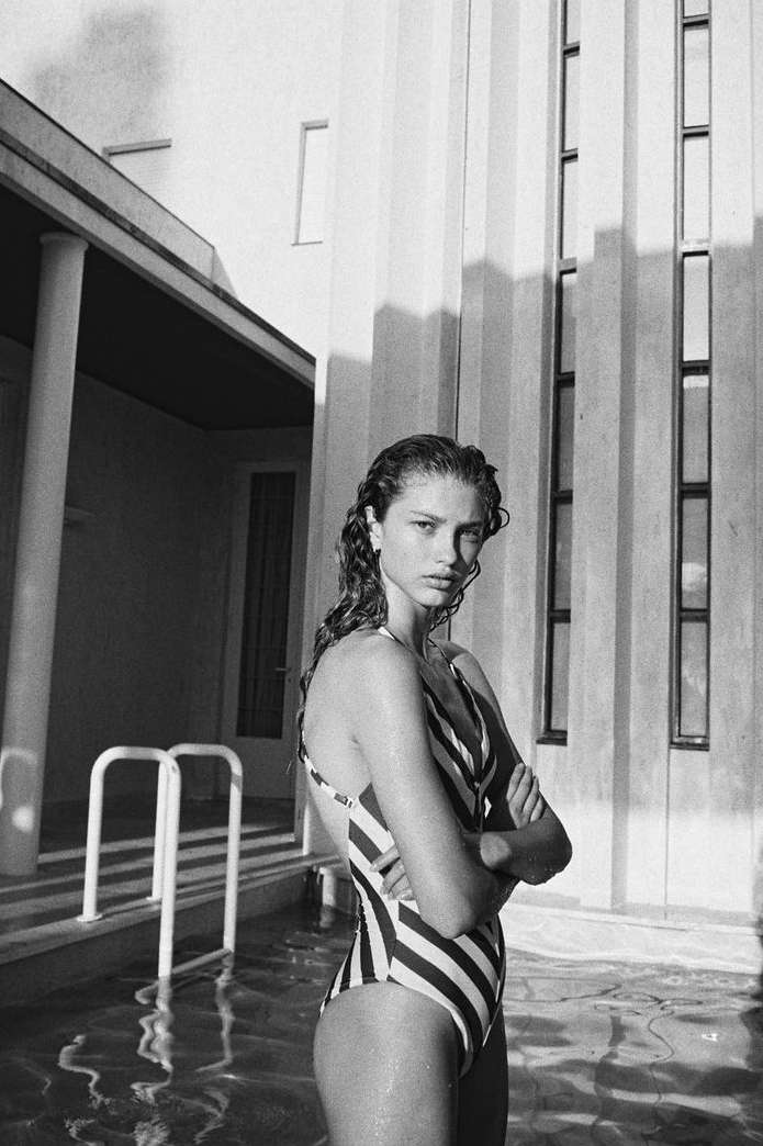 Woman in striped swimsuit stands in water near building with columns, wet hair, and crossed arms, portrait orientation.