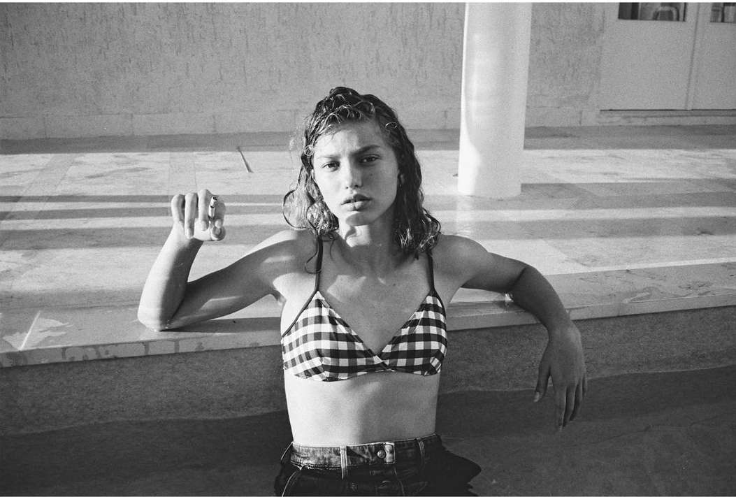 Monochrome portrait of a woman in a checkered bikini top and dark jeans leaning against a pool's edge, capturing a relaxed outdoor mood.
