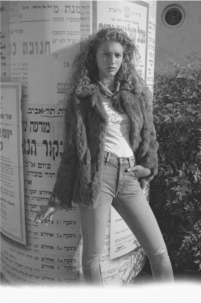 Black and white portrait of a stylish woman in a fur coat, jeans, and t-shirt, leaning against a pillar with posters outdoors.