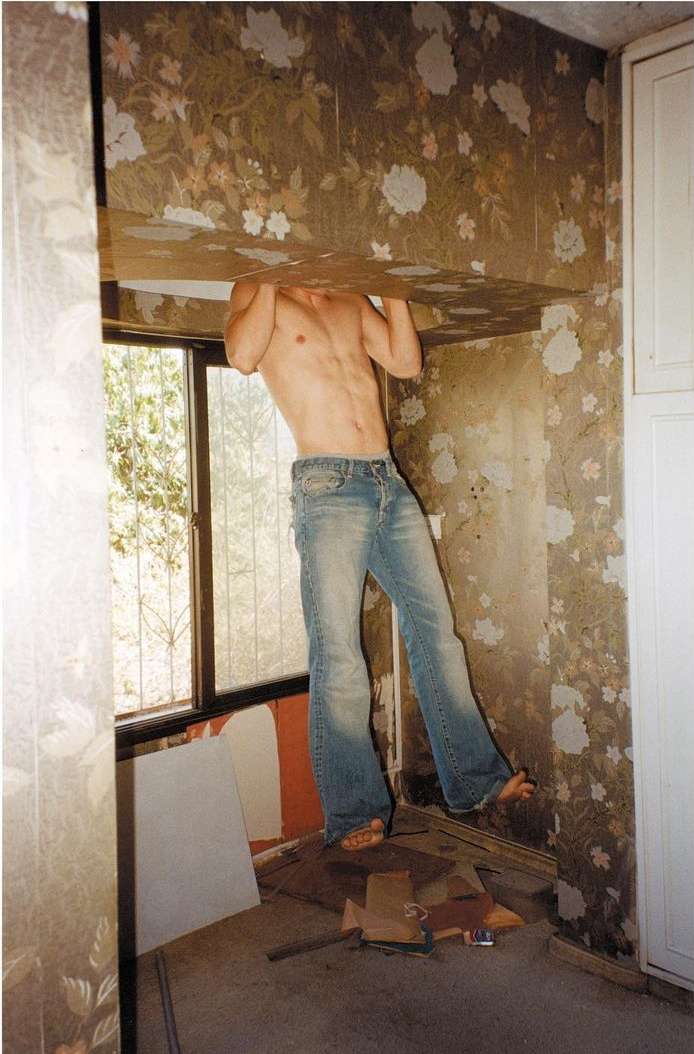 Man in jeans balances between a ceiling and floor with floral wallpaper in an empty room, near a window with natural light.