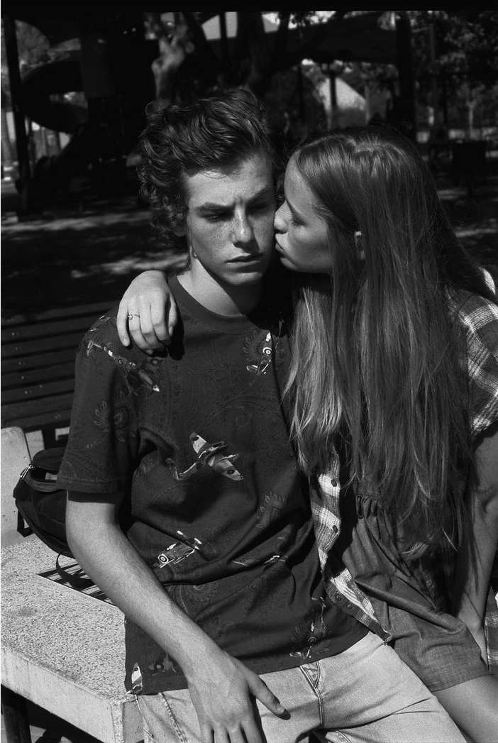 Black and white portrait of a young adult couple in a park-like setting; the woman kisses the man on the cheek in an outdoor setting.