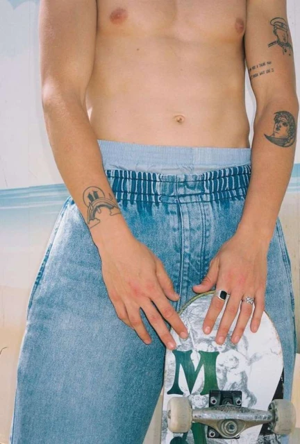 A shirtless young adult holds a skateboard in front of a pale-colored wall. He wears light blue denim pants and has tattoos on his arms.