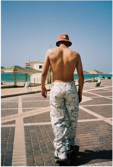 Back view of a shirtless male in patterned pants and bucket hat riding a skateboard near the ocean on a sunny day.