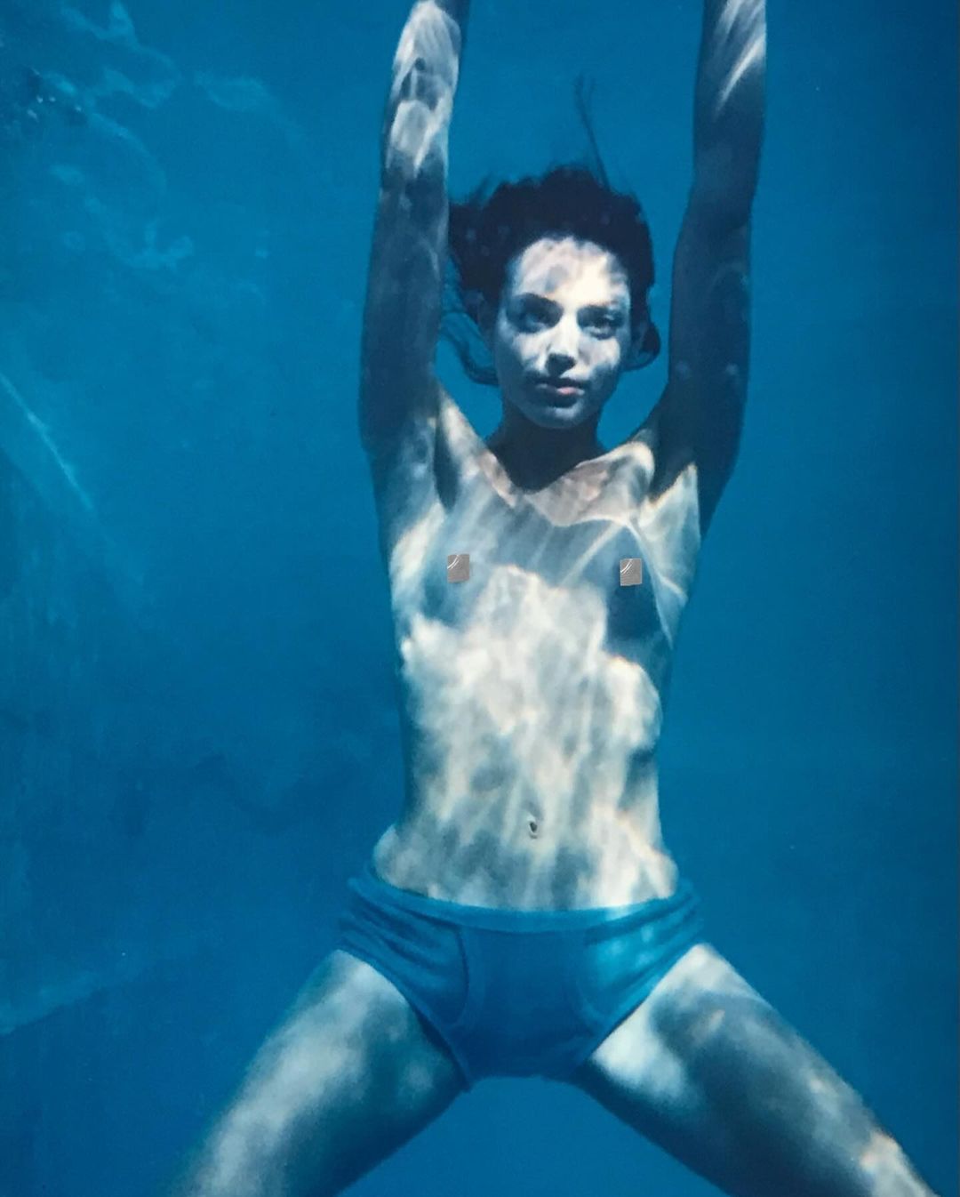 Underwater portrait of a woman with arms raised, wearing teal swimwear, surrounded by blue water with light patterns. 
