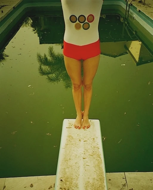 Woman in retro swimwear stands on weathered diving board above green pool, ready to dive in outdoor setting on a sunny day.