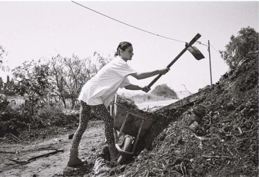 Black and white photograph of a woman digging with a shovel next to a wheelbarrow and a compost pile outdoors.