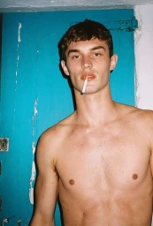 Shirtless young man with dark hair holding a cigarette in his mouth against a bright blue painted wooden door.