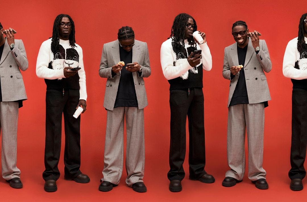 Three adult men in fashionable blazers and trousers pose on a red background, each holding or eating a snack while using a smartphone.