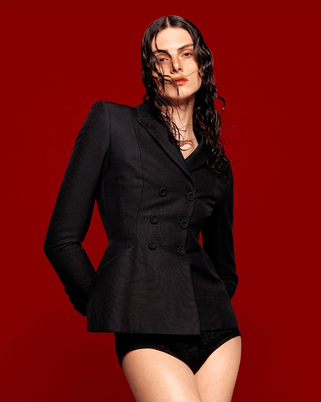 Adult female fashion model posing in a black double-breasted suit jacket and black briefs against a solid red studio backdrop.