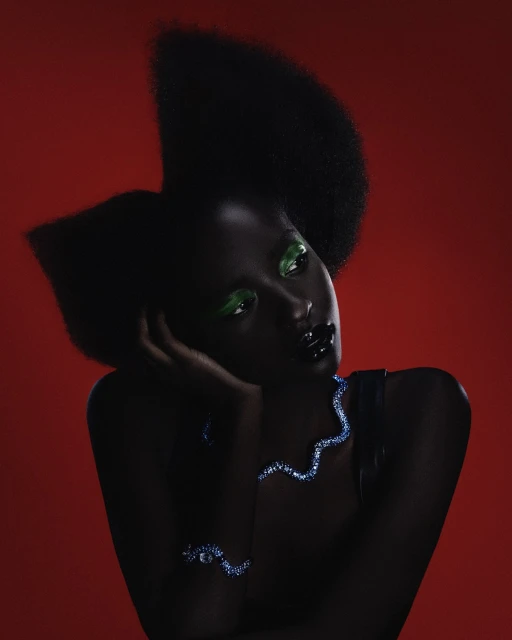 Portrait of a black woman with green eye makeup and afro hairstyle against a vibrant red backdrop, wearing a blue jeweled necklace and bracelet.