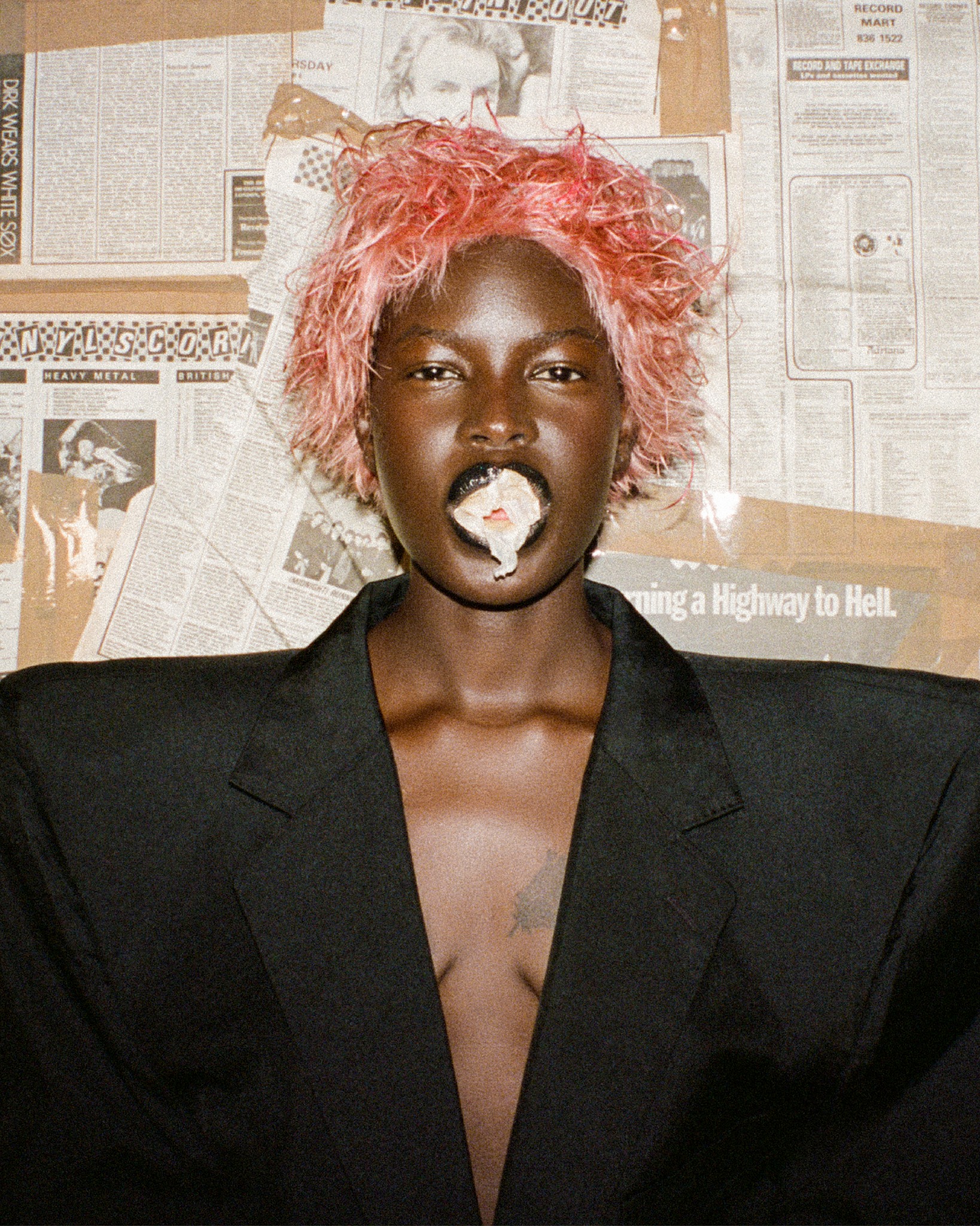 Portrait of a black woman with pink hair, wearing a black jacket, with white substance coming out of her mouth, posing against a newspaper background.