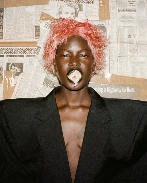 Portrait of a black woman with pink hair, wearing a black jacket, with white substance coming out of her mouth, posing against a newspaper background.