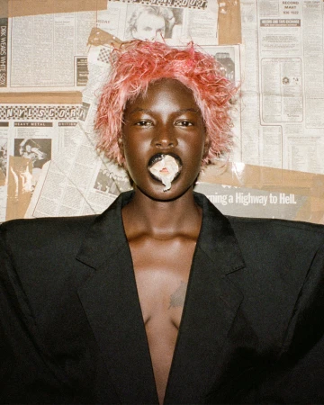 Portrait of a black woman with pink hair, wearing a black jacket, with white substance coming out of her mouth, posing against a newspaper background.