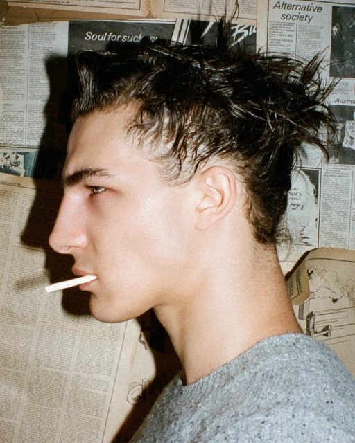 Profile of a young adult male with dark hair and a cigarette, posing in front of vintage newspaper clippings in soft, diffused lighting.