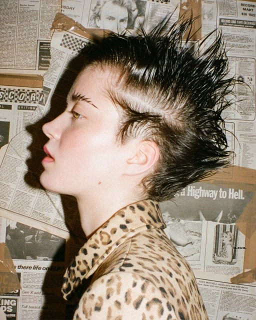 Profile of a female model with a wet-look spiky hairstyle and animal print jacket against a newspaper collage background in a studio.