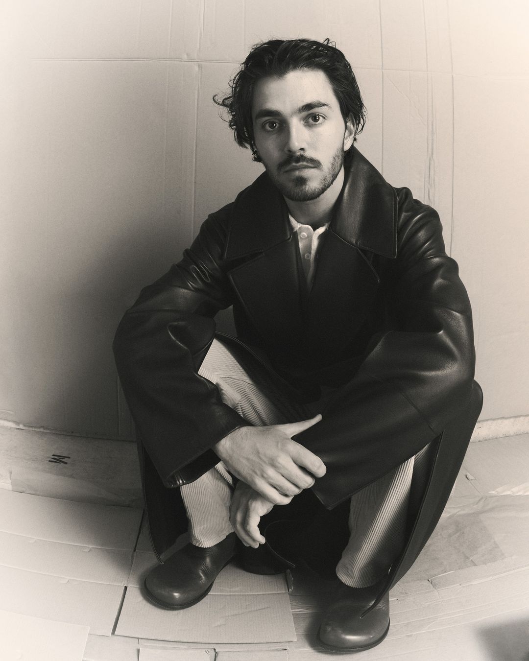 Portrait of a young adult man with a beard, wearing a black coat and boots, sitting in a photo studio with a cardboard background.