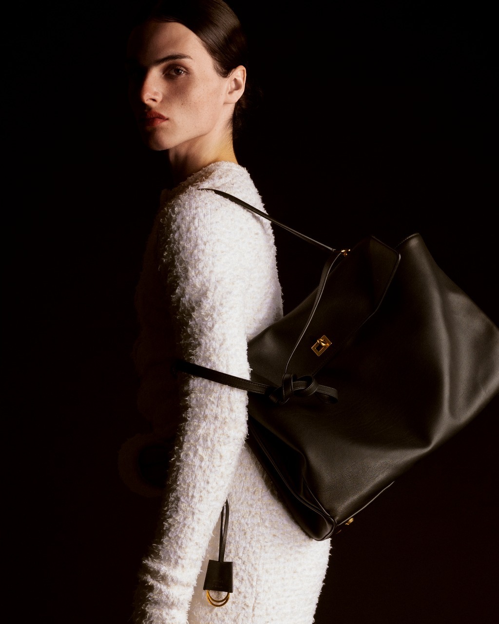 A woman with dark hair poses in a white textured dress with a large black leather tote bag on her shoulder against a dark backdrop.