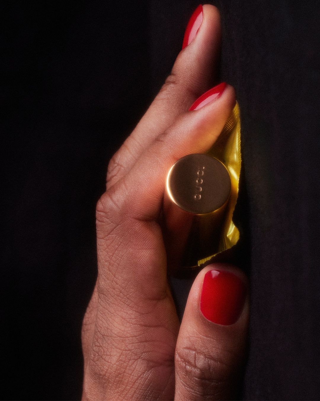 Close-up of a hand holding a golden tube of Gucci lipstick against a black background, showcasing luxury beauty.