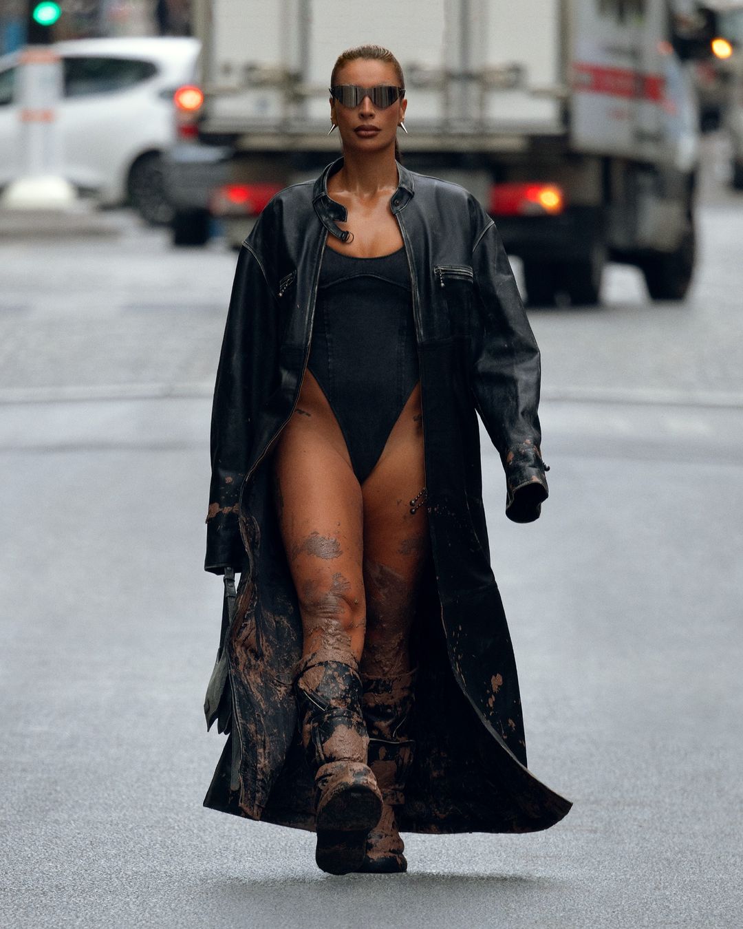 A woman walks on a city street wearing a black bodysuit, sunglasses, long coat, and boots. A white car and truck are behind her.