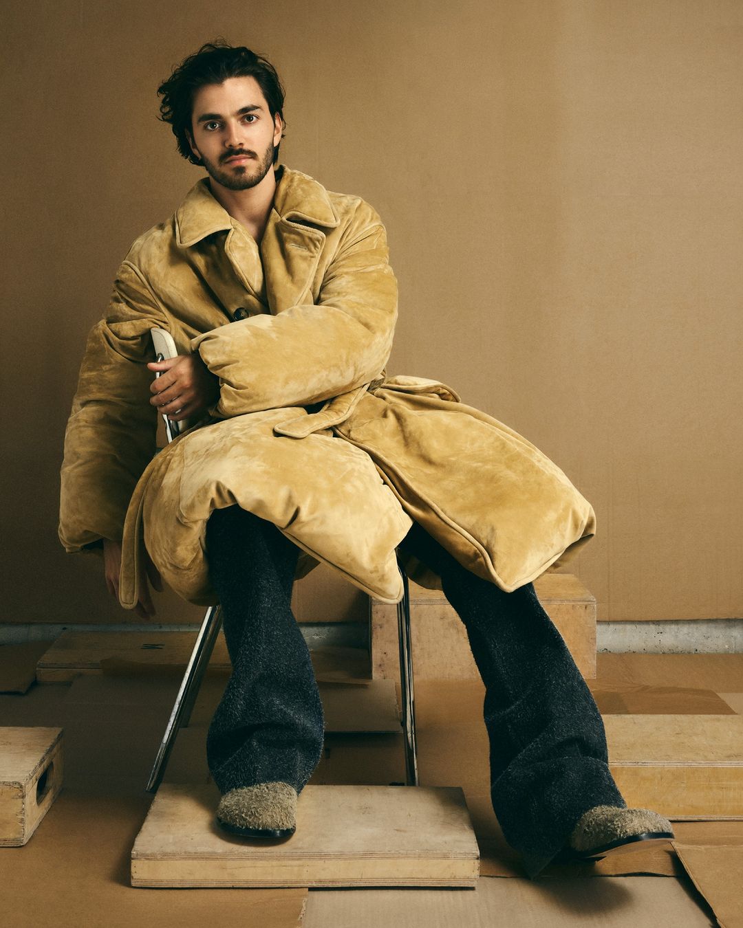 Man in tan coat sitting on chair in photo studio. The portrait captures modern fashion and neutral color palette in comfortable clothing.