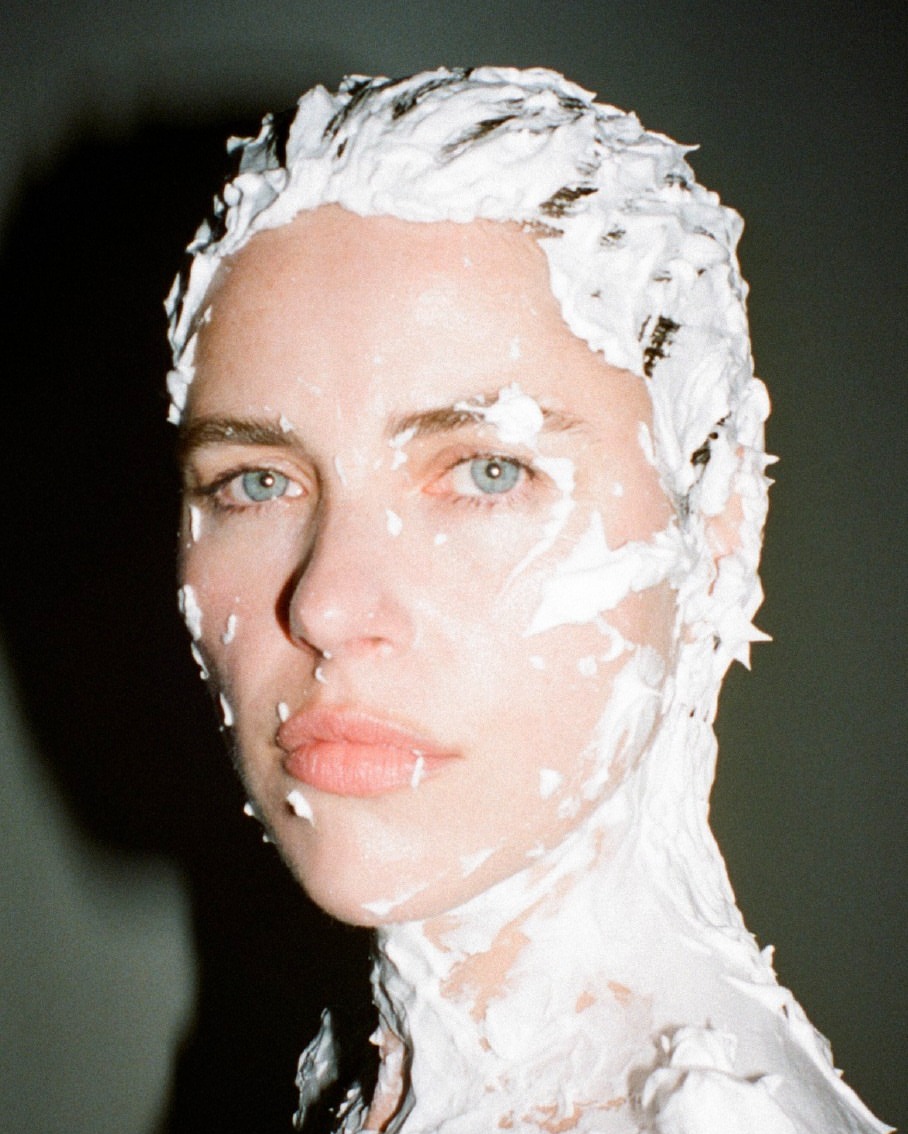 Portrait of a fair-skinned woman with blue eyes, covered in white creamy substance, captured in a close-up against a grey background.
