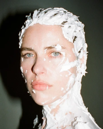 Portrait of a fair-skinned woman with blue eyes, covered in white creamy substance, captured in a close-up against a grey background.