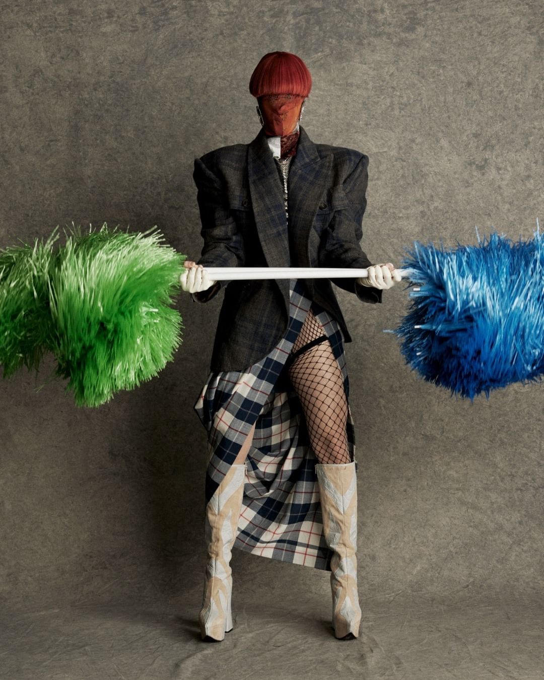 Person wearing an oversized plaid blazer, skirt, fishnets, and boots, holds a white bar with green and blue pompoms in a studio shot.